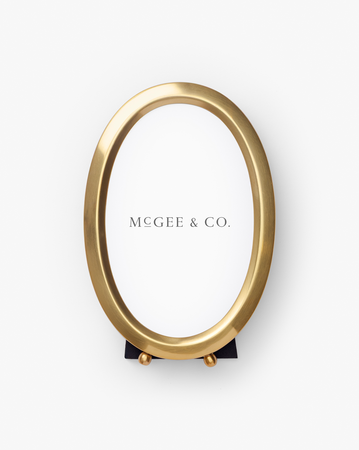 The Blanche Picture Frame is an oval, elegant photo frame with a polished brass finish, a white insert displaying "McGee & Co.," and a small black stand at the bottom, all shown on a plain white background, by Vendixs