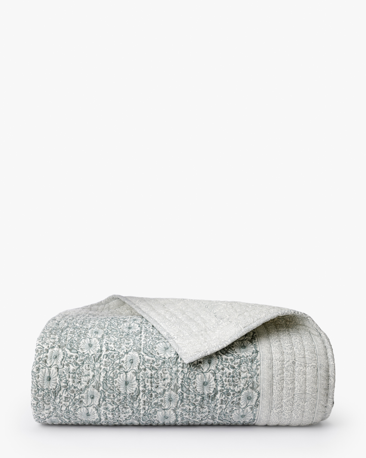 The Blue Floral Block Print Quilt, featuring a neatly folded design with a textured light gray and white pattern, is displayed on a plain white background. A turned-over corner reveals the artisanal bedding’s reverse side, by McGee & Co.