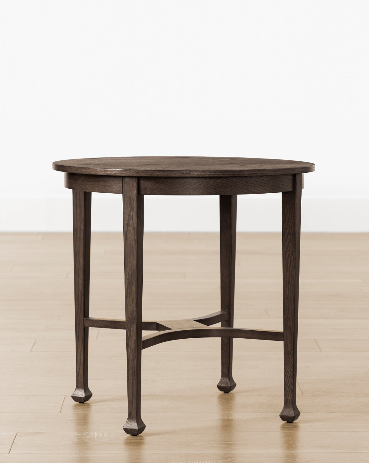 The Bradshaw Side Table showcases a round, dark satin walnut oak finish with four legs and a crossbar support, displayed on a light wood floor against a plain white wall, by Vendixs