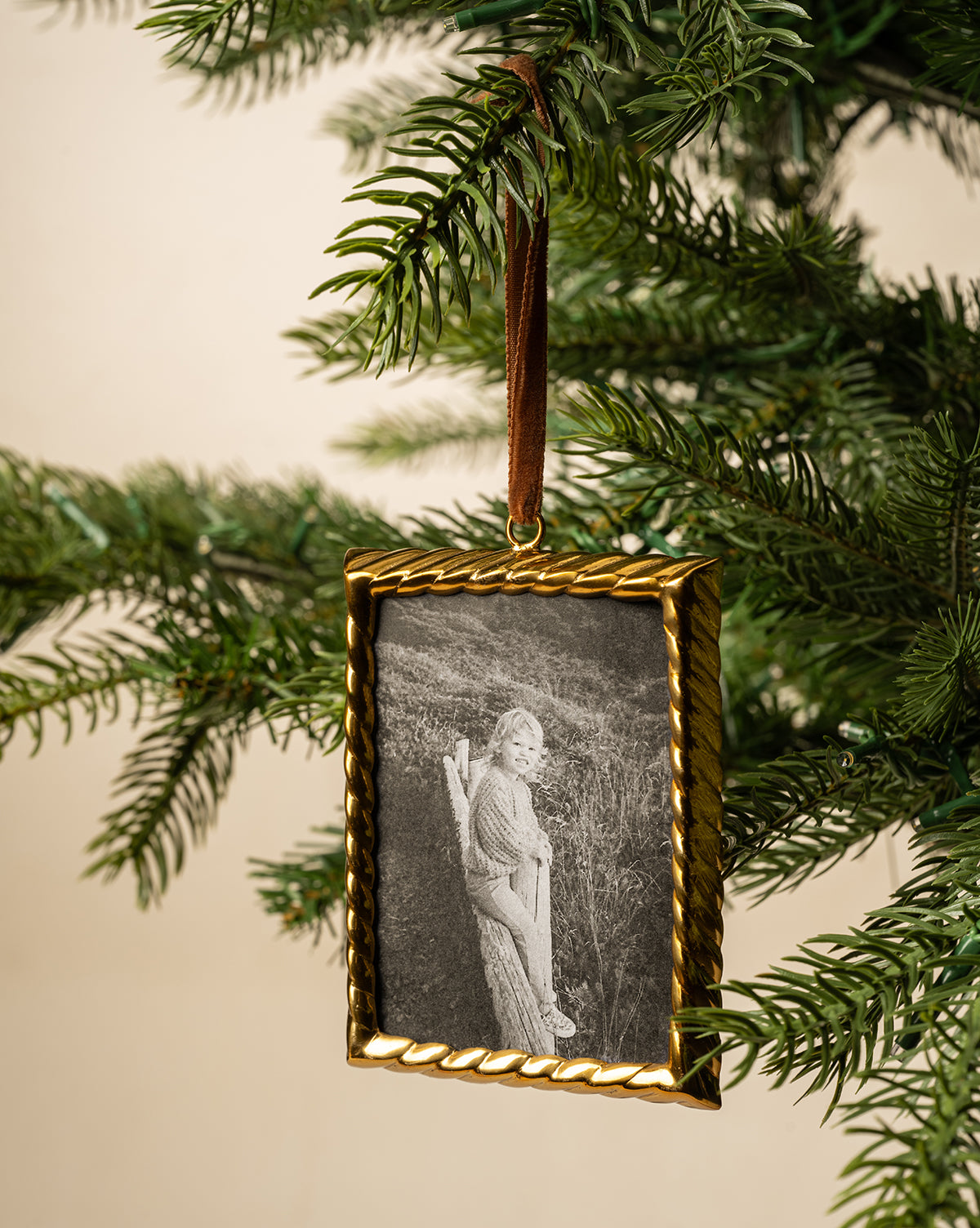 A Braided Brass Frame Ornament featuring a black-and-white photo of a child hangs from a brown ribbon on a green pine tree branch, by Vendixs