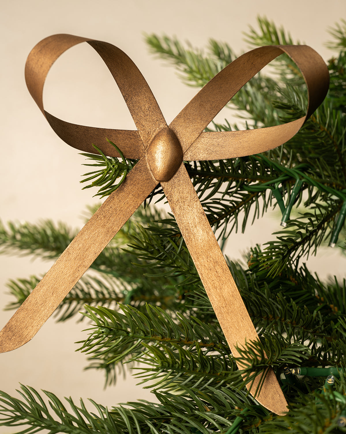 A Brass Bow Clip Ornament, featuring a metallic gold ribbon bow, is attached atop a green pine branch, offering a simple yet elegant touch against a neutral backdrop—ideal for adding festive charm to any Christmas tree, by Vendixs