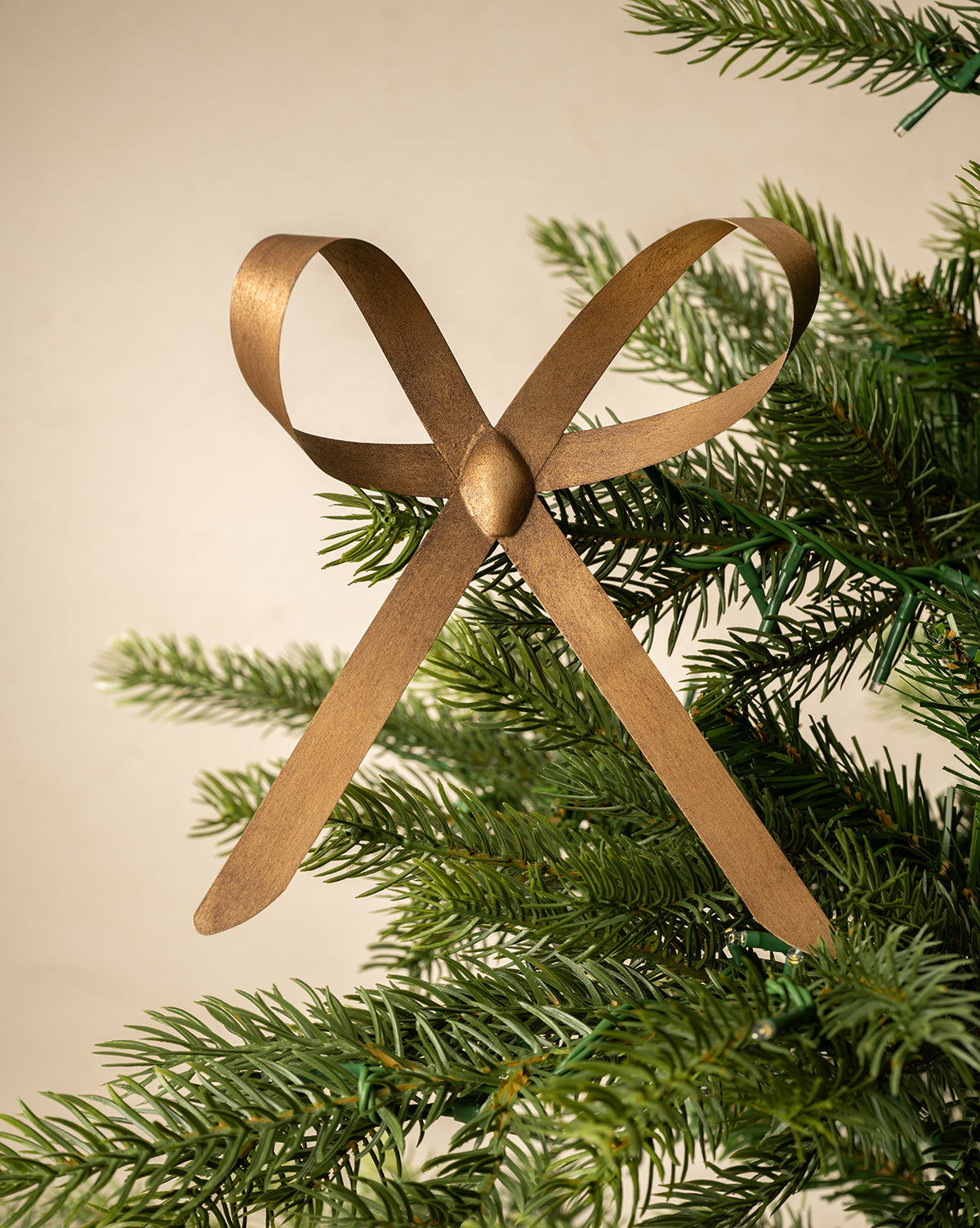 The Brass Bow Clip Ornament, featuring an antique brass finish, is clipped to the branch of a green Christmas tree against a plain beige background, by Vendixs