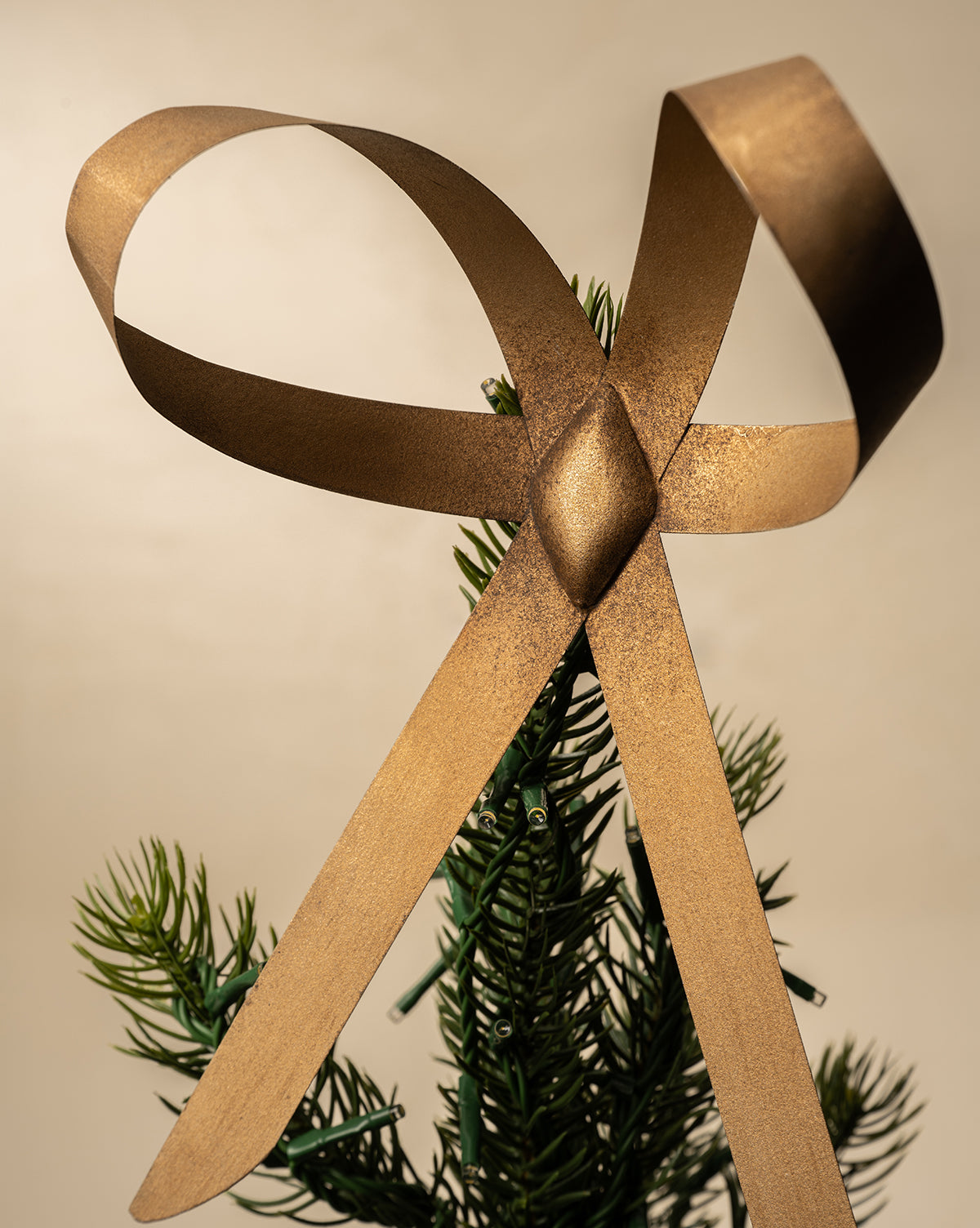 The Brass Bow Tree Topper, featuring an antique brass finish, sits in a classic bow atop the green tip of a Christmas tree against a neutral background, by Vendixs