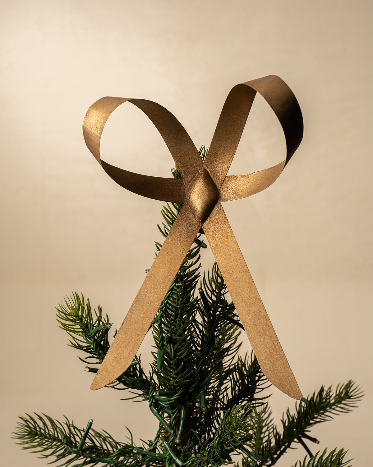 The Brass Bow Tree Topper, featuring an antique brass finish, is placed atop a Christmas tree’s green branches with a plain beige background, by Vendixs