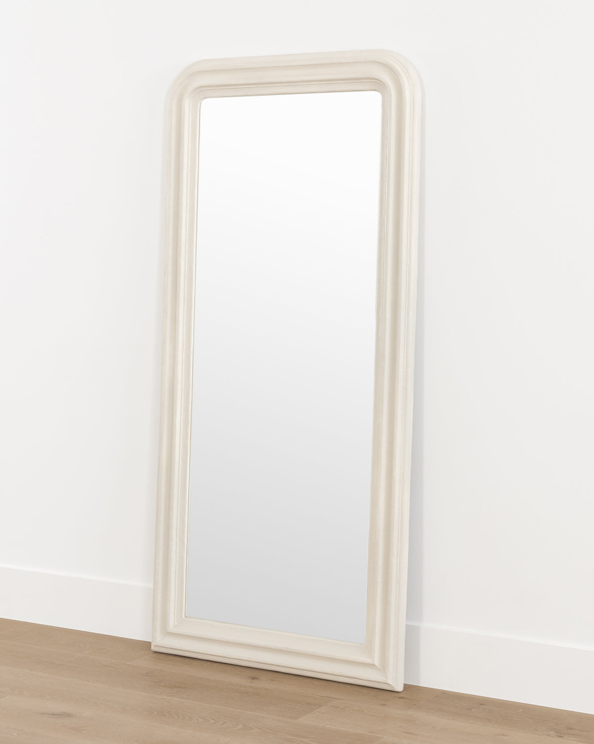 The Breslin Floor Mirror, featuring a tall, rectangular shape with a rounded top and a thick off-white plaster finish frame, leans against a white wall on a light wood floor, by Vendixs