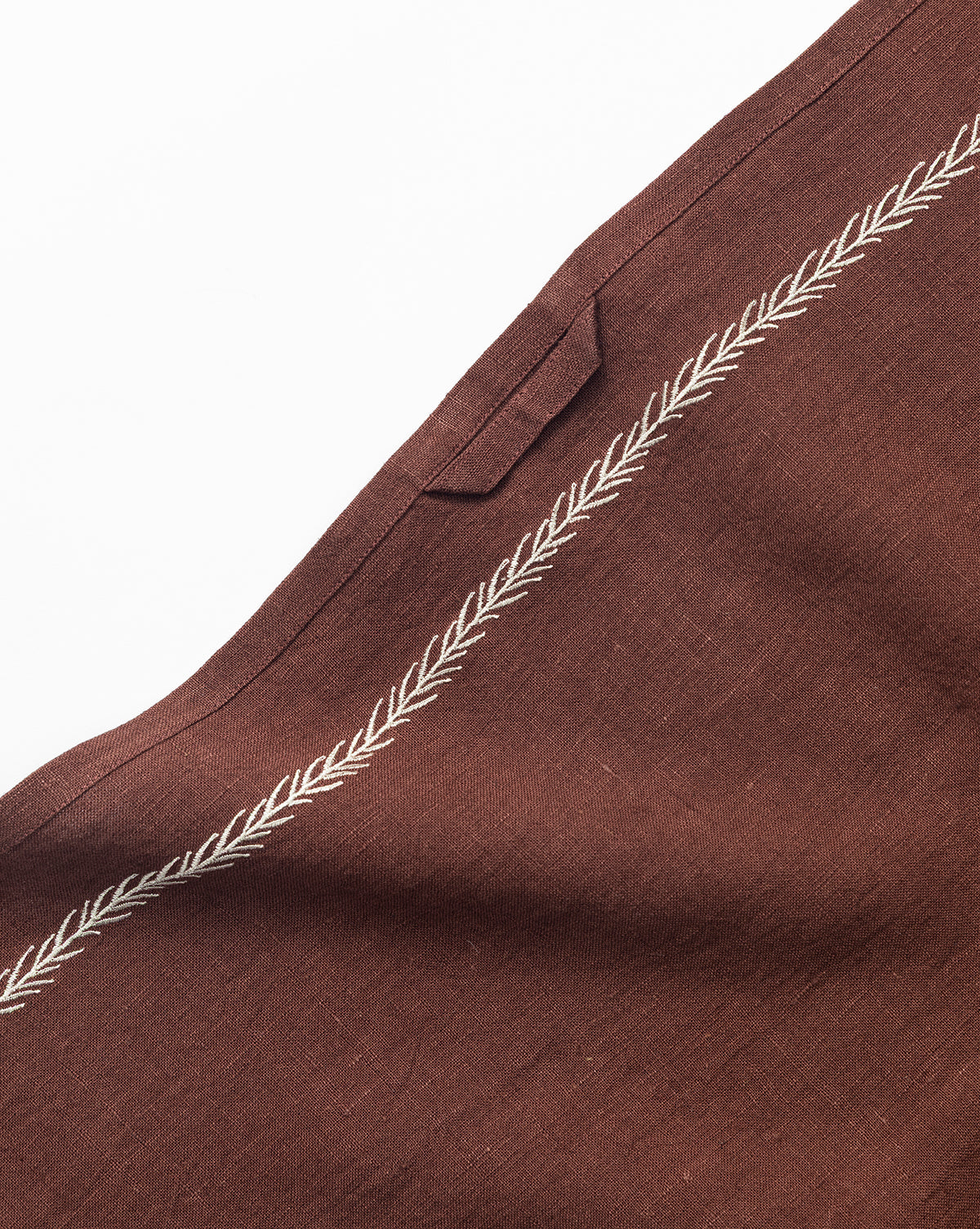 A close-up of the Broadleaf Tea Towel, featuring a brown fabric with an embroidered leaf border and a small rectangular loop—an elegant kitchen accessory set against a white background, by Vendixs
