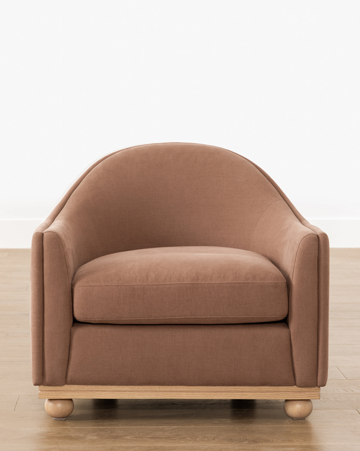 The Bunnell Lounge Chair (Ready to Ship) by Makerspalm features brown upholstery, a rounded backrest, low wide seat, and light wooden base with round feet, shown on a wooden floor against a white wall. Vendixs