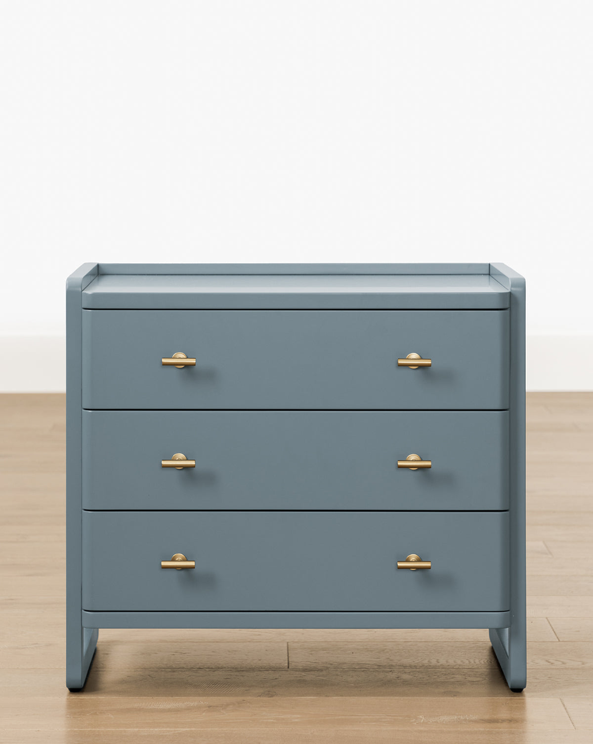 The Butler Nightstand features a modern slate blue finish, three drawers with gold handles, and stands on a light wooden floor against a plain white background, by Vendixs