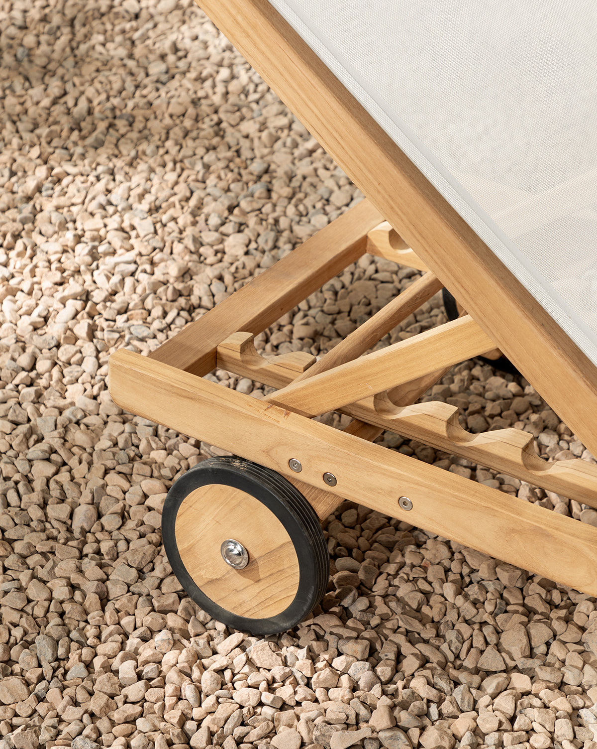 Close-up of the Cadie Outdoor Chaises teak frame and wheel on light pebbles. Featuring a weather-resistant mesh seat and an adjustable backrest, this chaise is designed for outdoor comfort by McGee & Co.