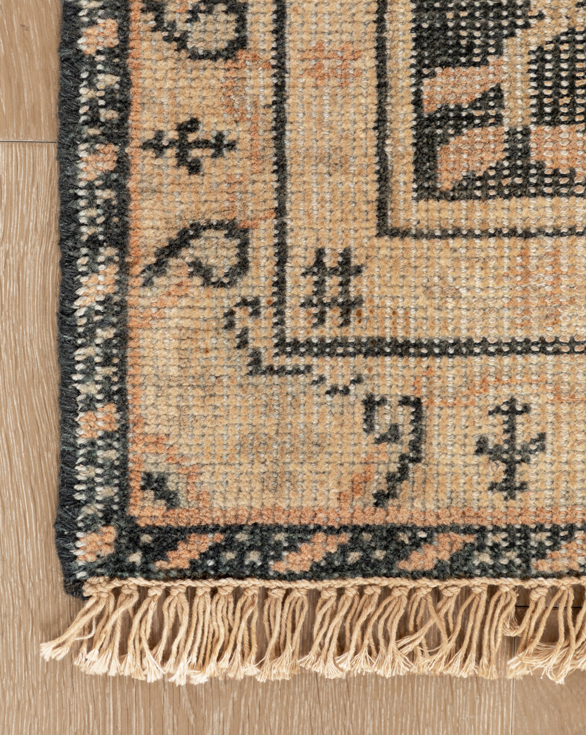 Close-up of the Campanella Hand-Knotted Wool Rug, showcasing a geometric border in beige, black, and peach tones with artisan fringe details on the edge, displayed on a wooden floor, by Vendixs