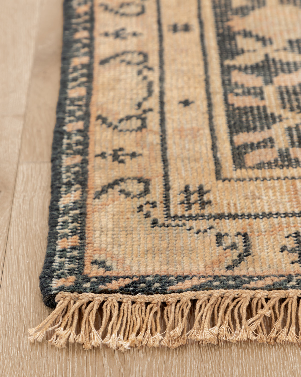 Close-up of the Campanella Hand-Knotted Wool Rug, showcasing intricate geometric patterns in beige and dark gray, artisan details, and edge fringe on a light wooden floor, by Vendixs