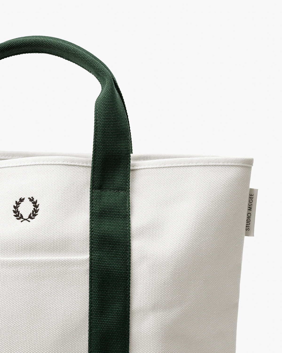 A close-up of the Canvas Laurel Tote, featuring a white canvas body, green handle, embroidered laurel wreath logo, and a small STUDIO NICHOLSON tag on the side, by Vendixs