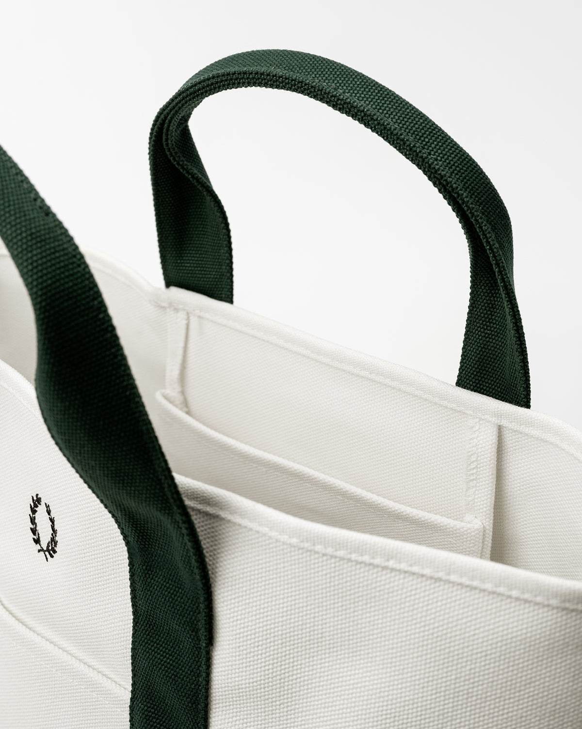A close-up of the Canvas Laurel Tote, an everyday bag with dark green handles and trim, an embroidered wreath logo, and visible inner pockets, shown against a plain white background, by Vendixs