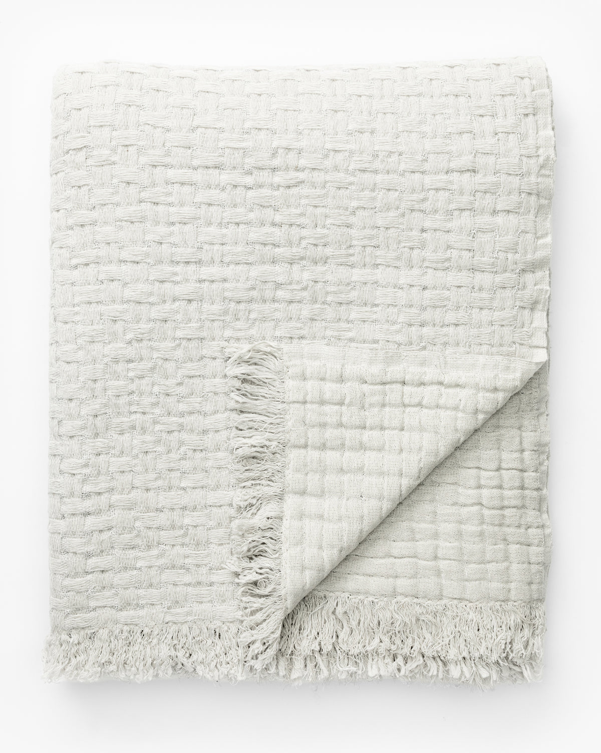 The Caroline Kids Blanket by Indigo Craft is a folded, textured off-white throw with a subtle checkered pattern and fringed edges, displayed on a white background with one corner flipped to show the reverse side. Vendixs