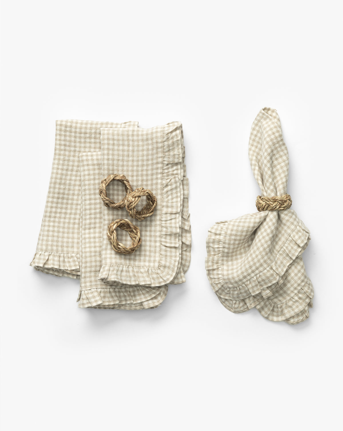 The Catania Linen Napkins (Set of 4) in beige and white gingham are styled with ruffled edges—three folded with braided straw napkin rings, and one elegantly unfolded—displayed against a white background, by Vendixs