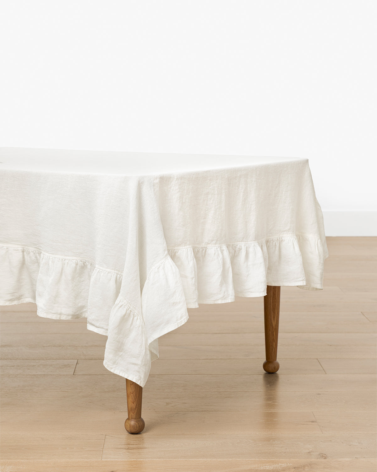 A rectangular wooden table with curved legs is draped in the Catania Linen Ruffle Tablecloth, by Vendixs The table stands on a light wood floor against a plain white wall.