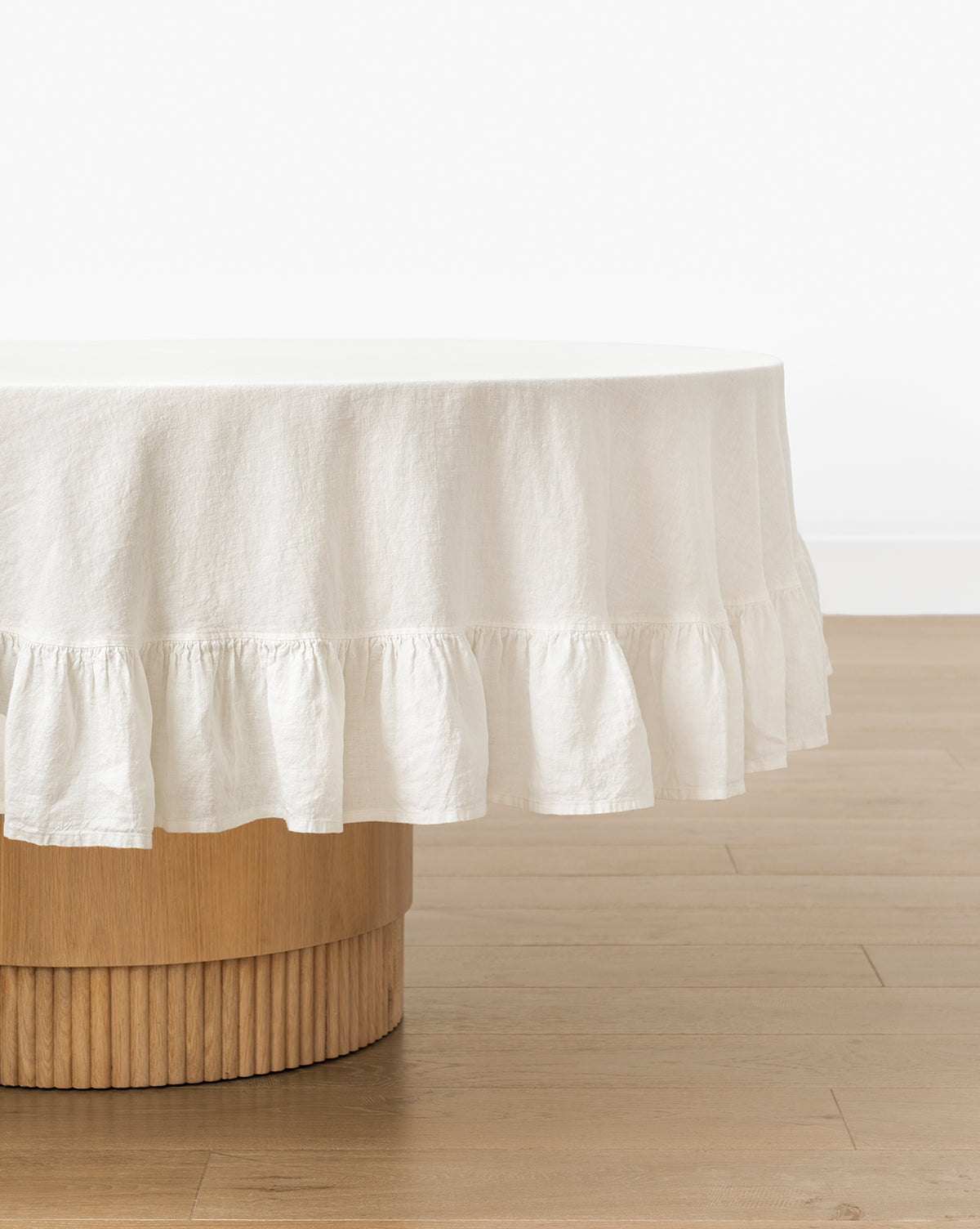 The Catania Linen Ruffle Tablecloth, with a delicate ruffled edge, covers a round wooden table with a ribbed base, displayed on a light wooden floor against a plain white wall, by Vendixs