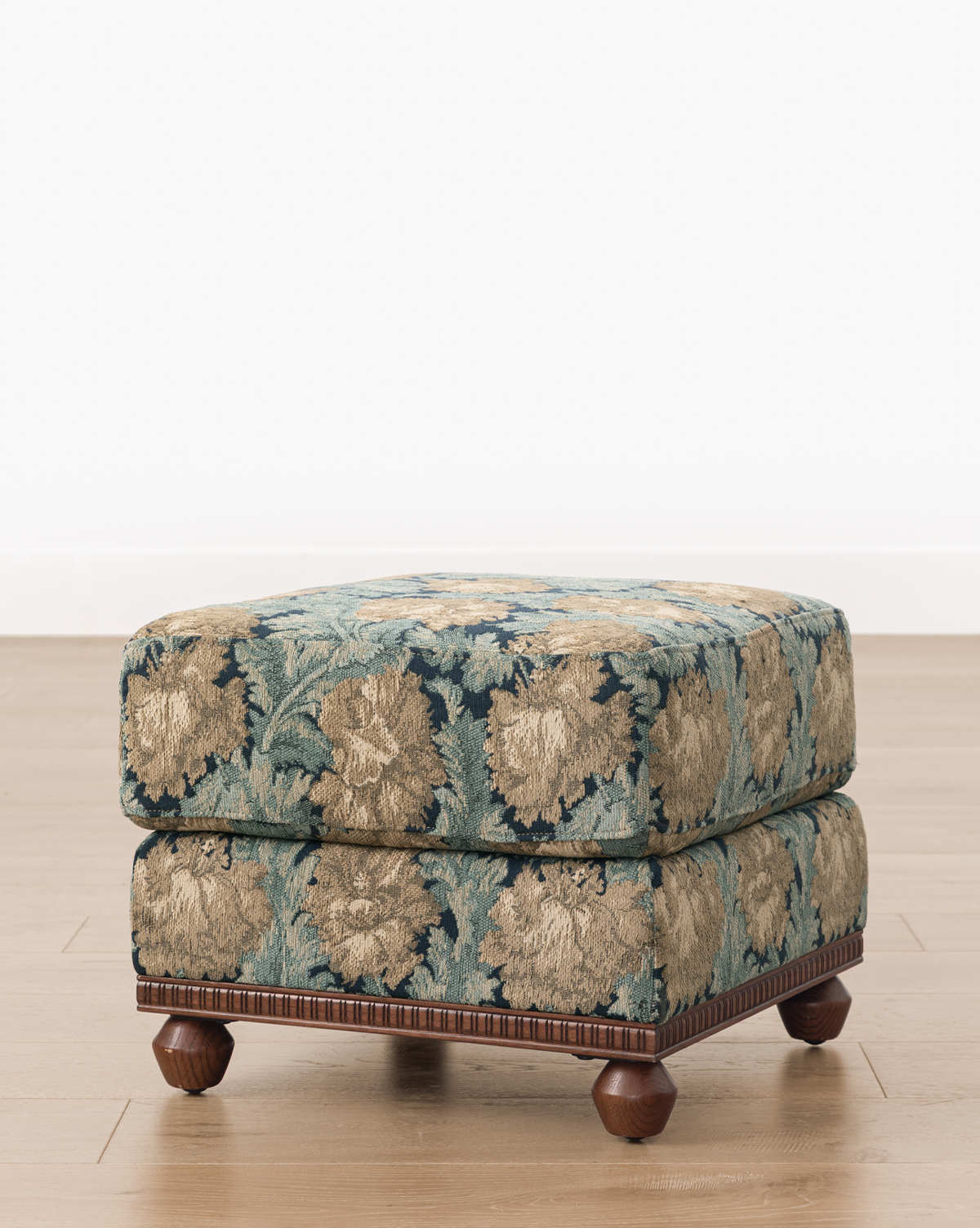 The Catherine Ottoman features a square, upholstered design with blue and beige floral fabric, a carved wood base, and round wooden feet, set on a light wood floor against a white background, by Vendixs