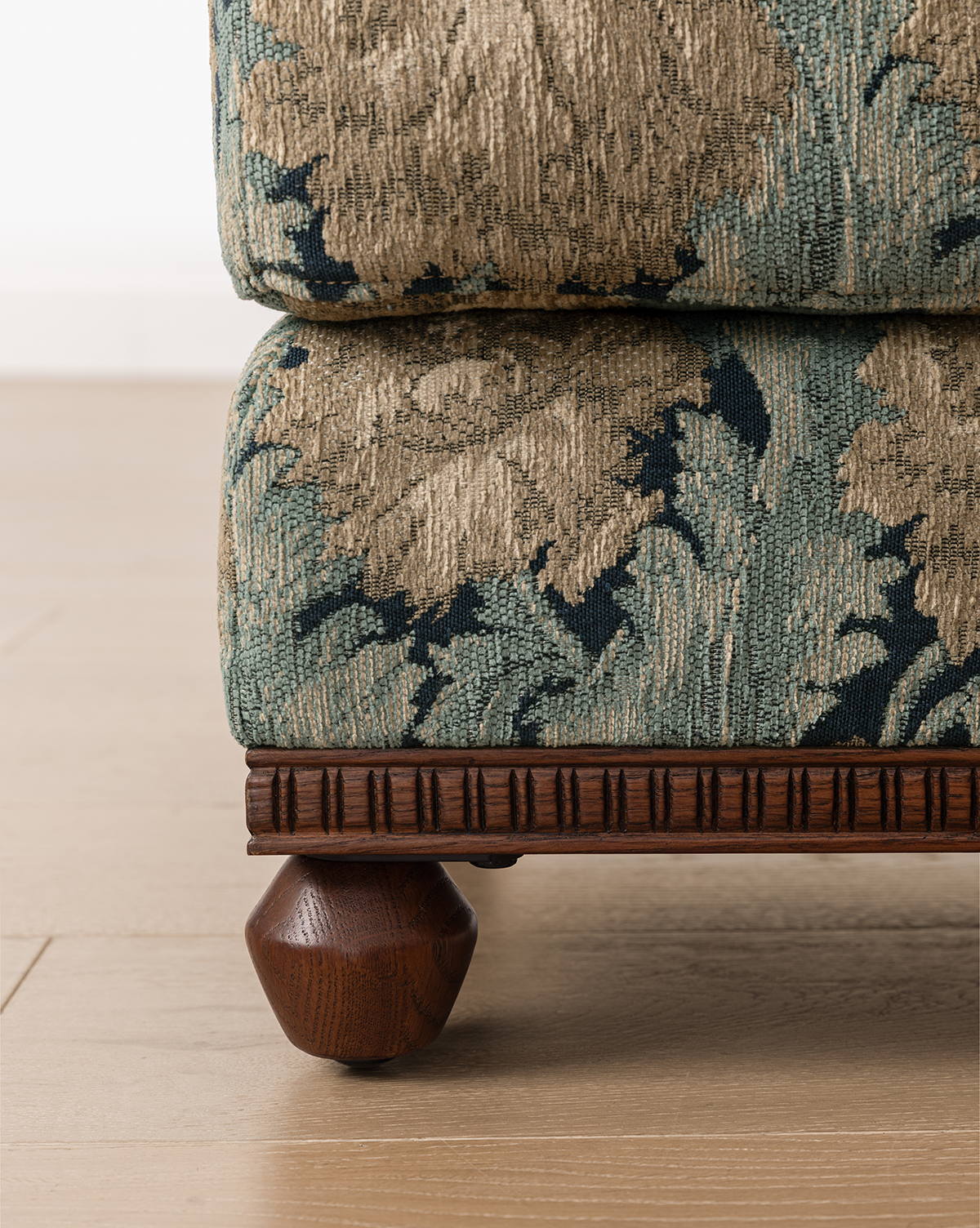 Close-up of the Catherine Ottoman, upholstered in green and beige patterned fabric, with a carved wood base and a round dark wooden leg, shown on a light wood floor, by Vendixs