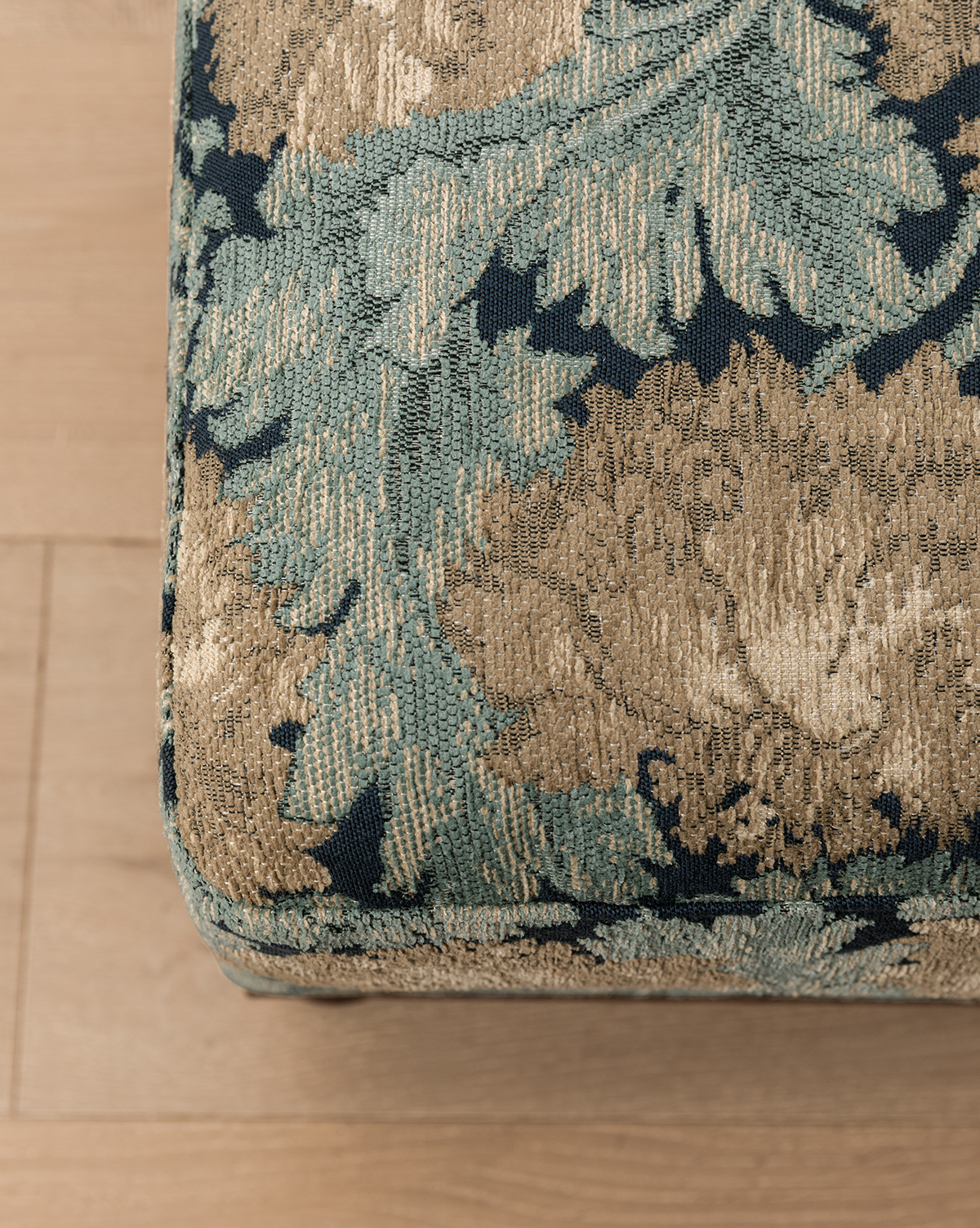 Close-up of the Catherine Ottoman corner, featuring green and gold floral patterned fabric with a carved wood base, on a light wooden floor, by Vendixs