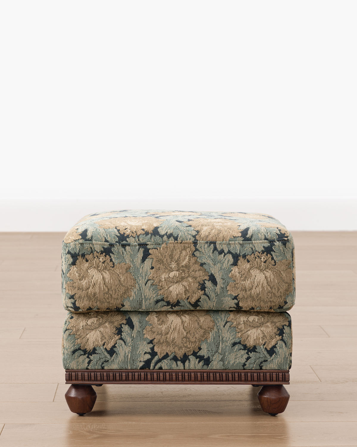 The Catherine Ottoman, featuring a carved wood base and ornate blue and beige floral upholstery, is placed on a light wood floor against a plain white wall, by Vendixs