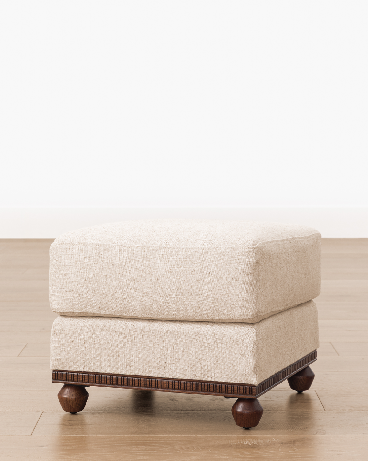 The Catherine Ottoman has a square beige upholstered top and dark carved wood base with four round wooden legs, adding elegance to any space, by Vendixs