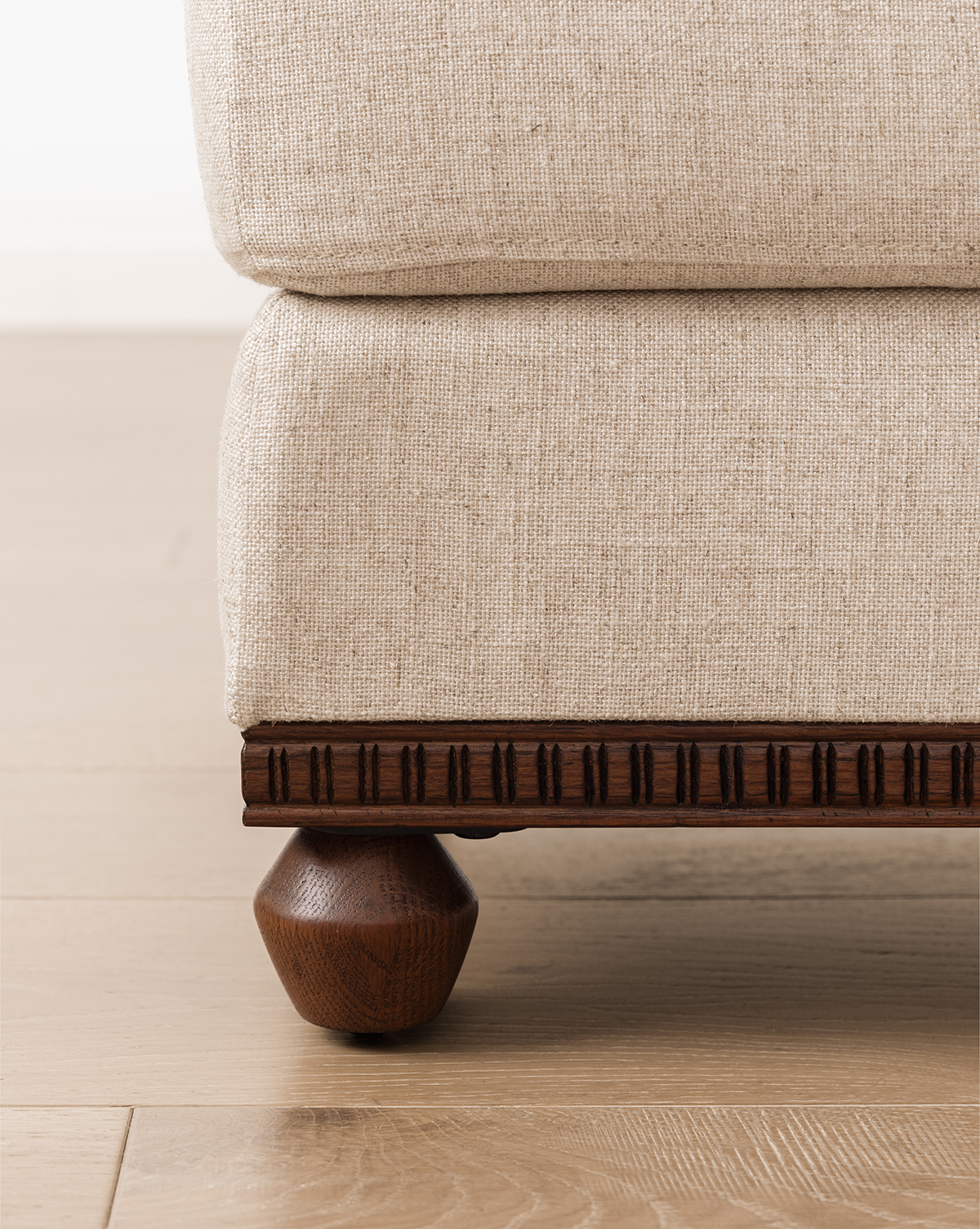 Close-up of a beige upholstered sofa with a carved wood base and round wooden leg, placed on a light wood floor beside the Catherine Ottoman, by Vendixs