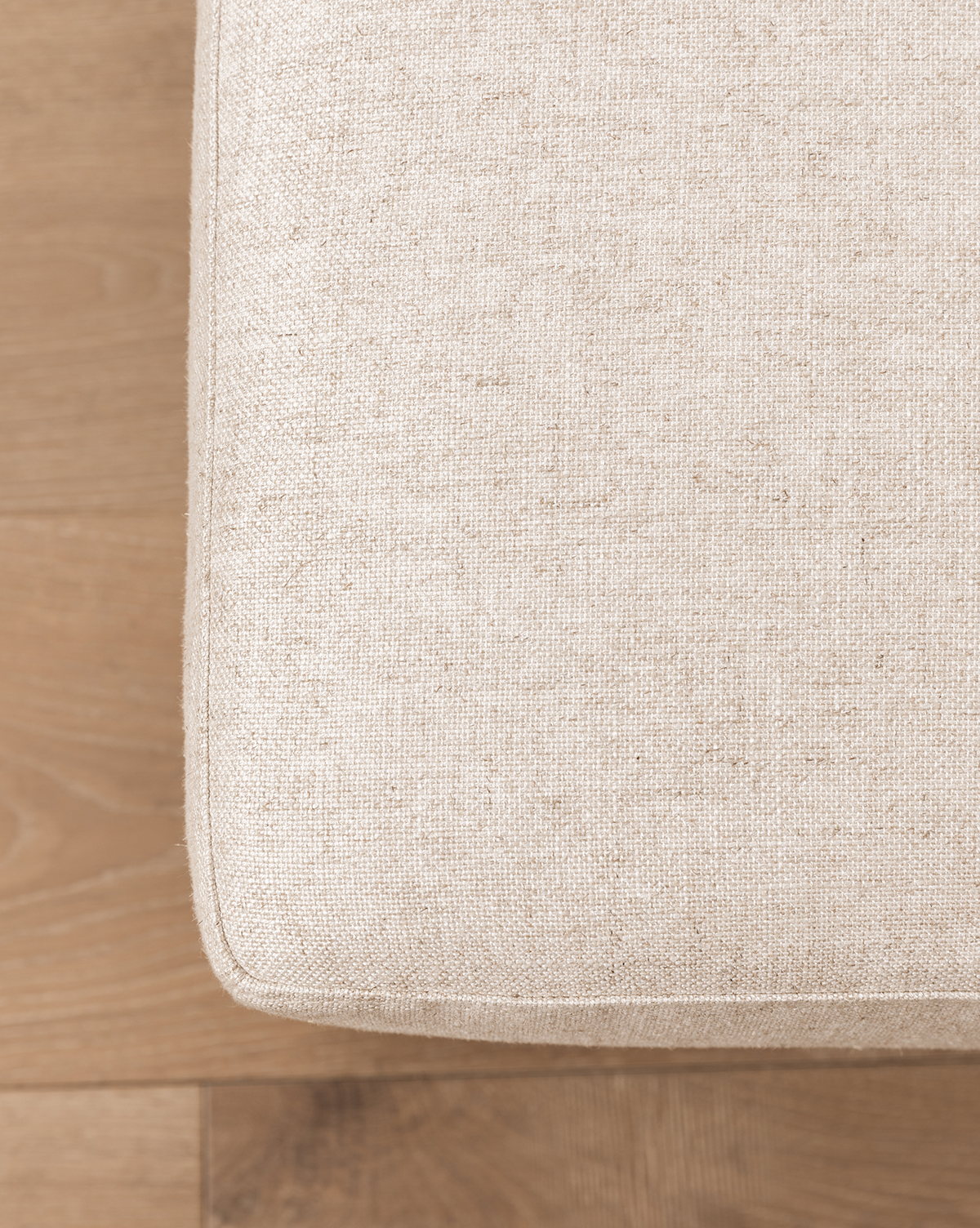 Close-up of the corner of a beige fabric cushion on the Catherine Ottoman, set on a light wooden floor. The upholstery texture and wood grain are clearly visible, by Vendixs