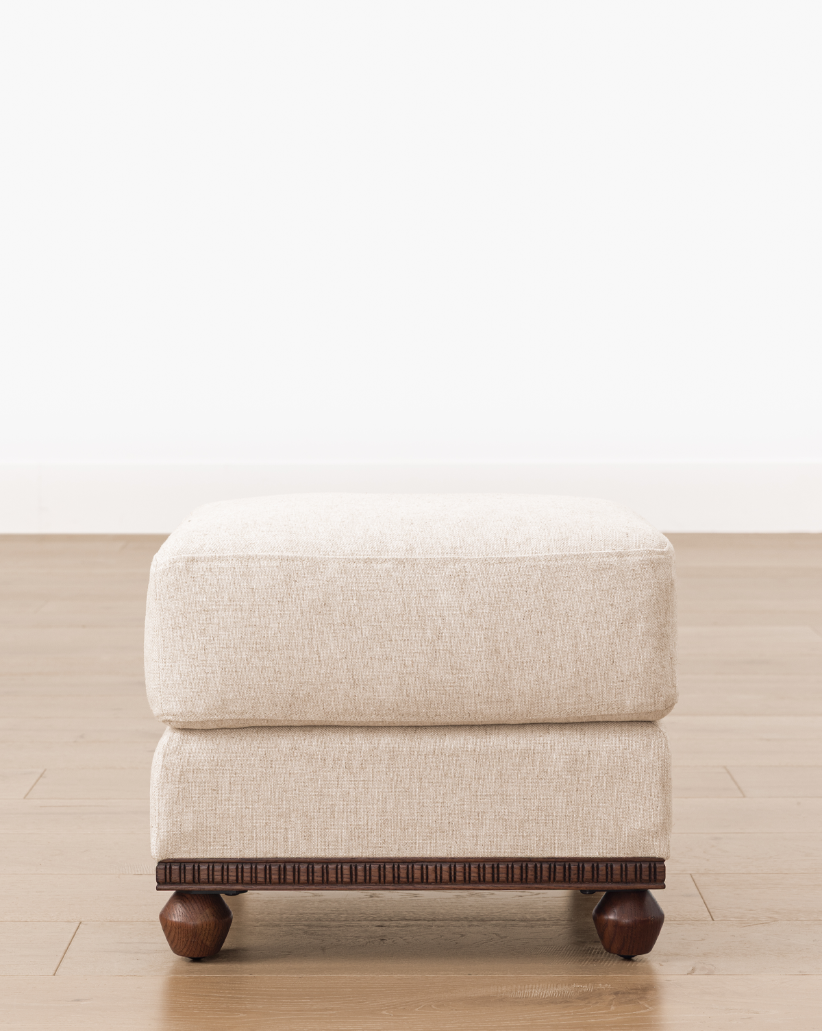 This upholstered ottoman looks stylish on a light wood floor beside a plain white wall, by Vendixs