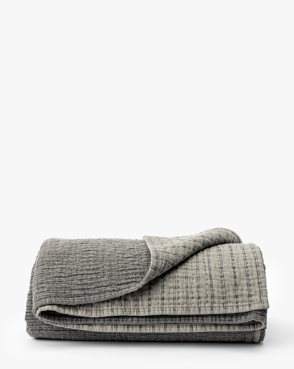 The Cecily Kids Quilt by Renuka Inc. is a textured gray blanket with a visible stitched pattern, neatly folded on a flat white surface. One corner is slightly flipped over to show the underside, Vendixs