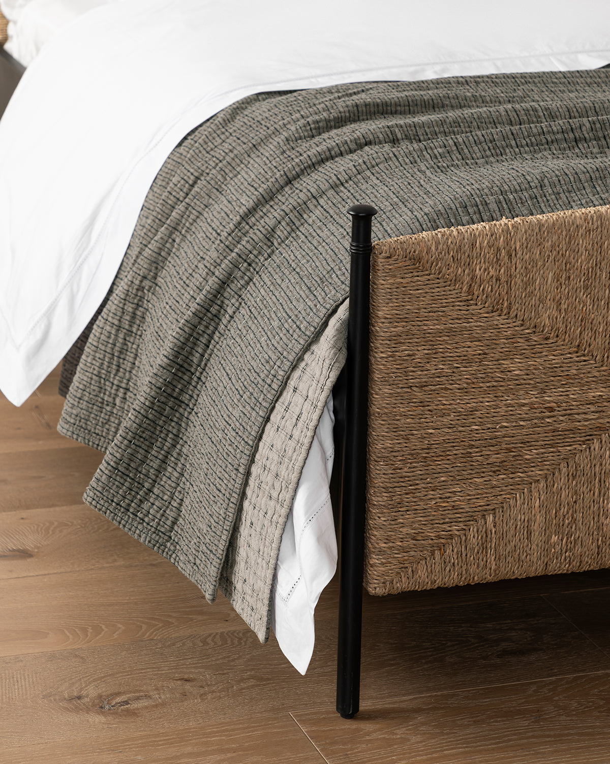 A close-up of a bed corner with a woven rattan frame, white sheet, Cecily Kids Quilt, gray textured blanket, and a patterned beige throw, all on a wooden floor by Vendixs