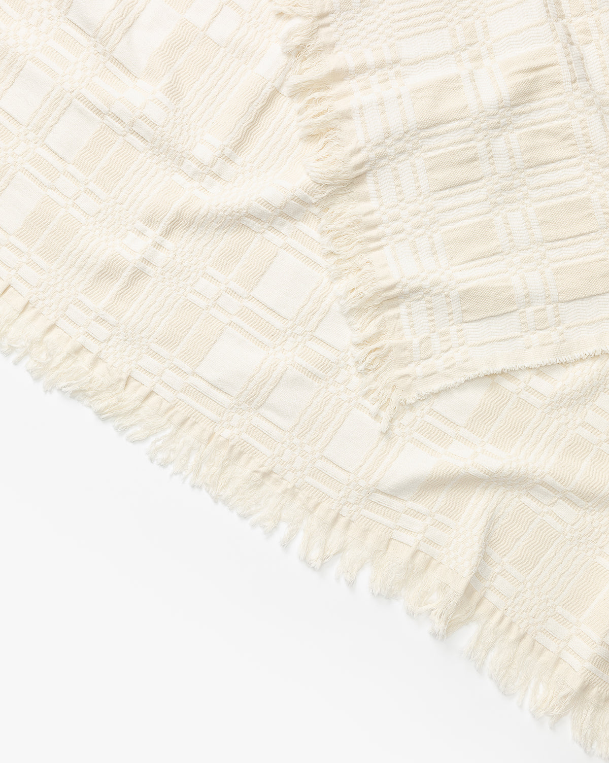 A close-up of the Chamberlain Coverlet by Makerspalm, featuring cream-colored cotton with a textured plaid pattern and fringed edges, arranged diagonally on a white background by Vendixs