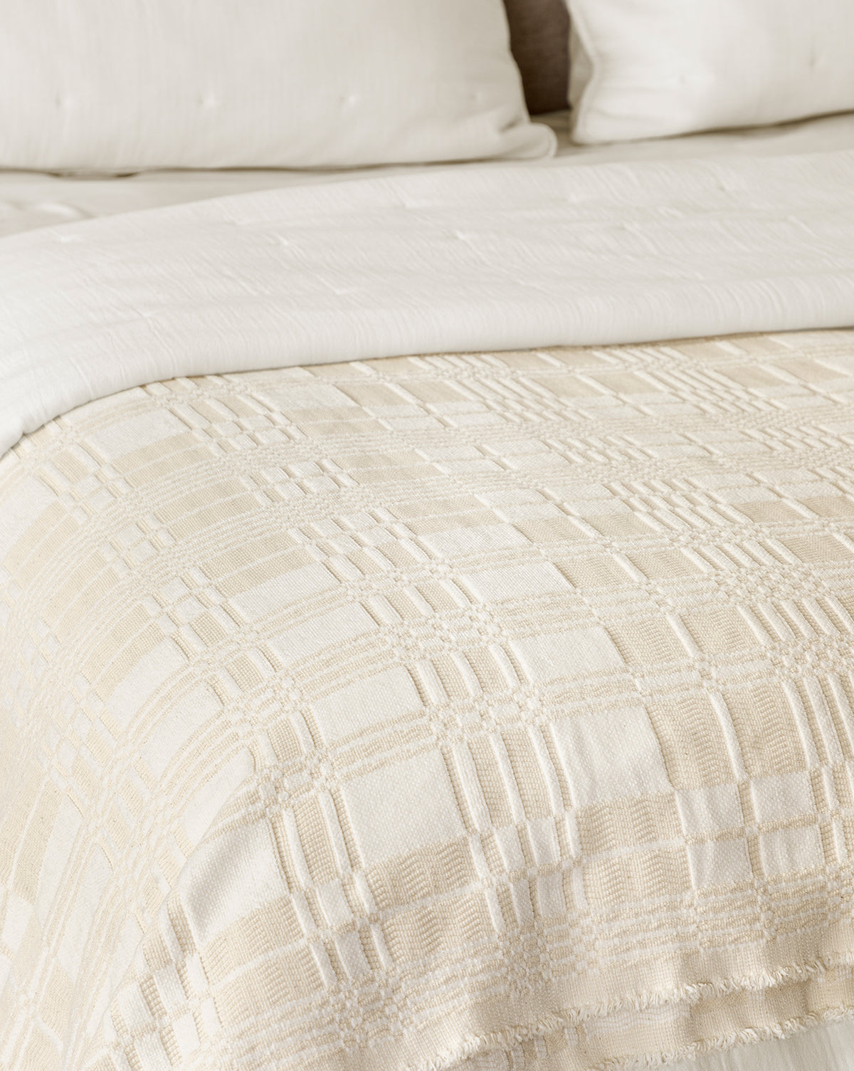 A close-up of a bed with cream bedding, showcasing the Makerspalm Chamberlain Coverlet, a textured cotton layer paired with a plaid woven blanket and matching pillows in the background, from Vendixs