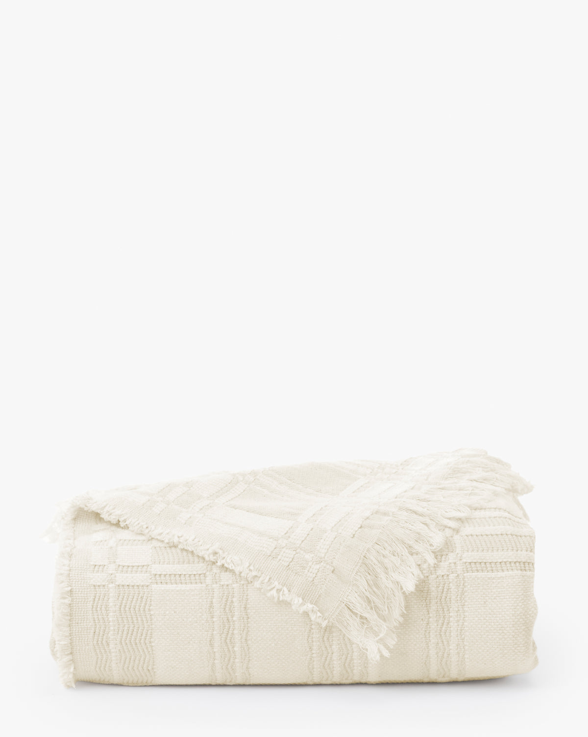 The Makerspalm Chamberlain Coverlet is a folded off-white woven blanket with a subtle geometric pattern and fringed edges, displayed on a plain white background. Vendixs