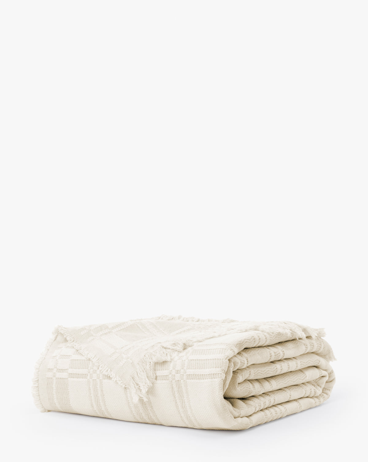 The Makerspalm Chamberlain Coverlet is a cream-colored woven blanket featuring a subtle checkered pattern and fringed edges, shown folded against a plain white background. Vendixs