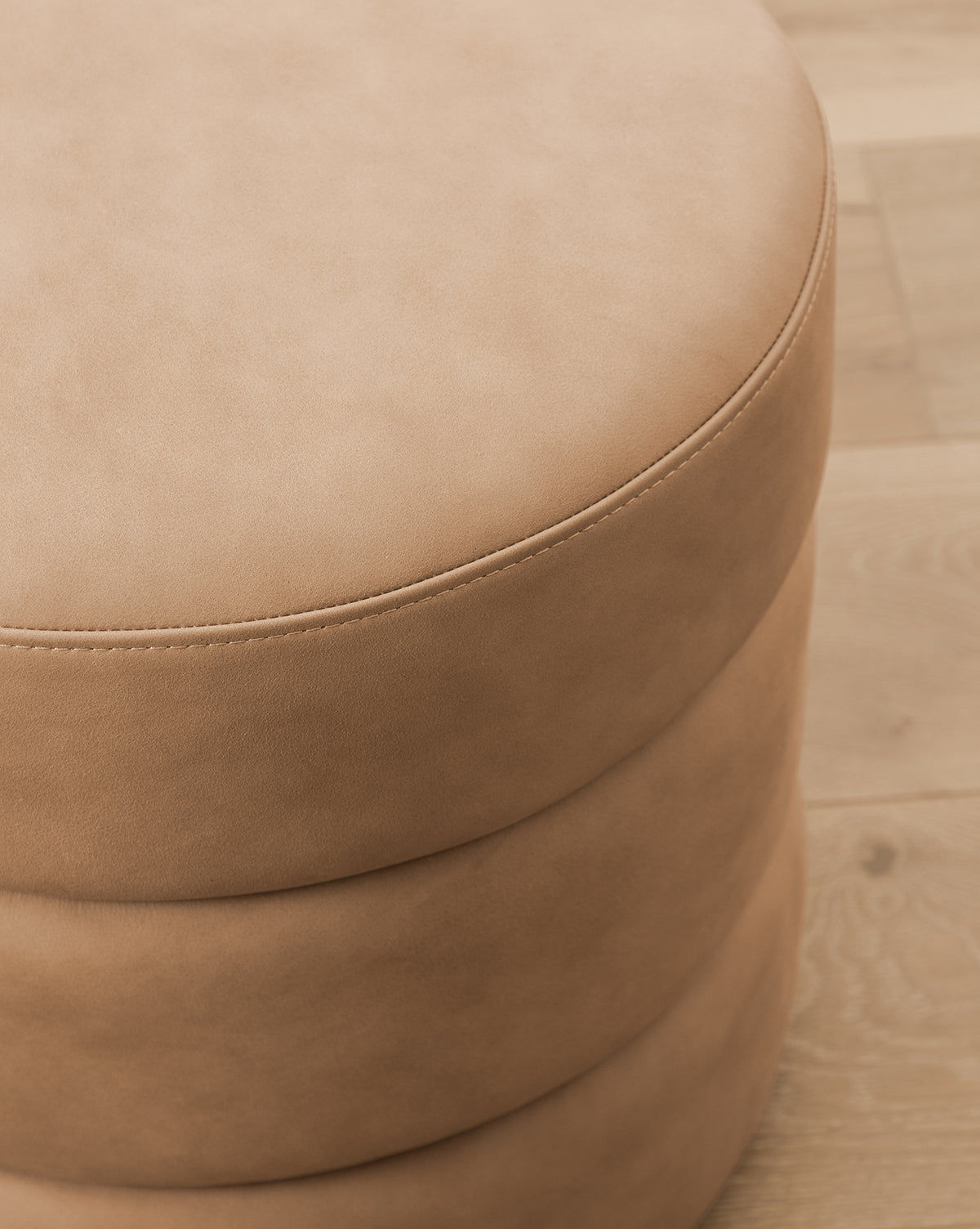 Close-up of the Channel Ottoman, a round, tan piece featuring visible top stitching and subtle quilted details, placed on a light wood floor to bring midcentury charm to any room, by McGee & Co.
