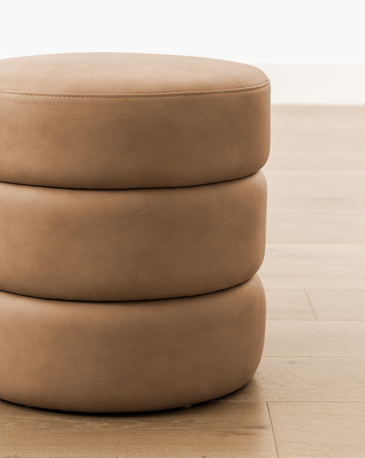 A close-up of the Channel Ottoman, a round, light brown, three-tiered piece with smooth upholstery, sits on a light wood floor against a white wall, bringing midcentury flair to the room by Vendixs