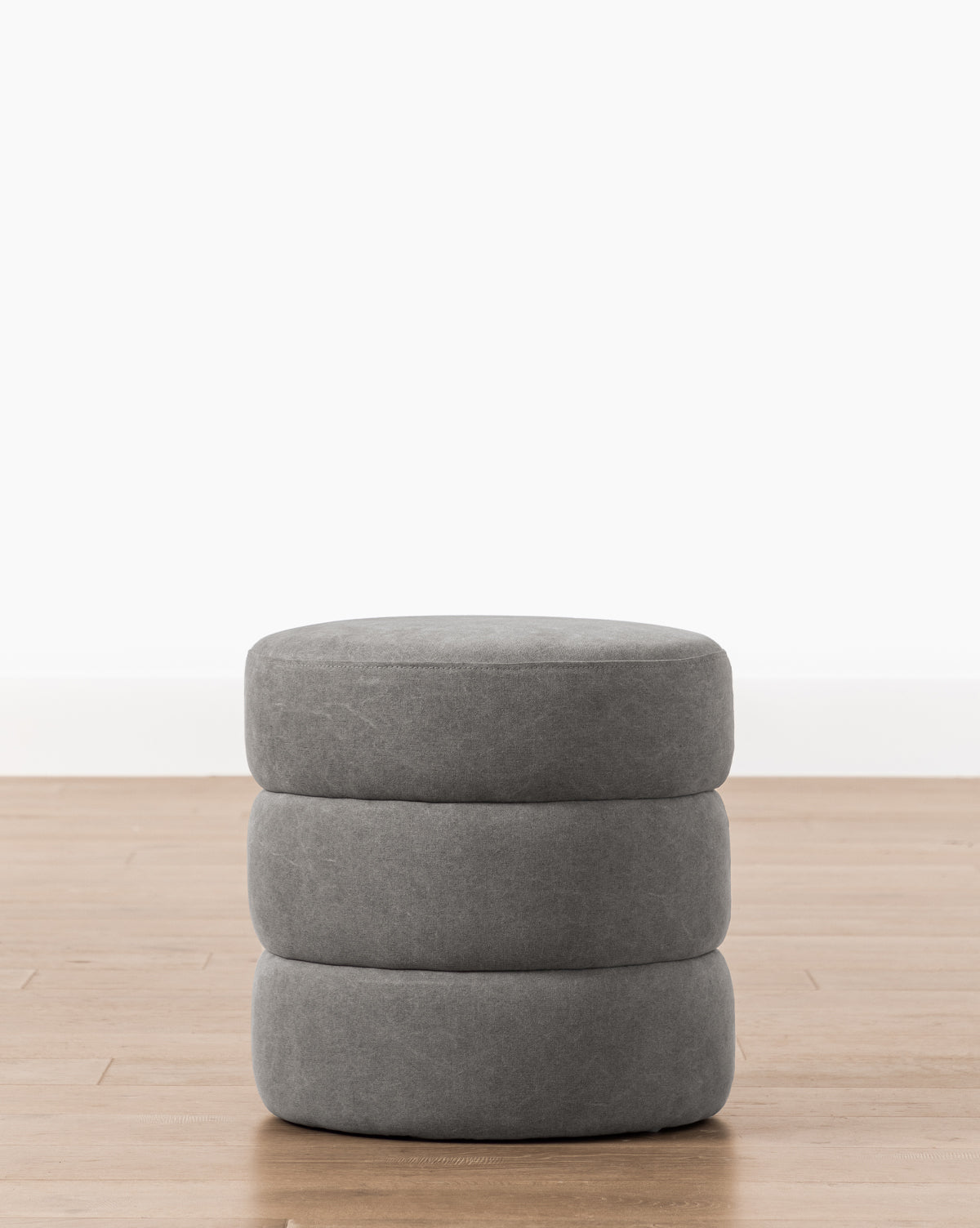 The Channel Ottoman features a round, gray design with three stacked cushion-like layers and a subtle quilted texture, adding midcentury charm as it sits on a light wood floor against a plain white wall, by Vendixs