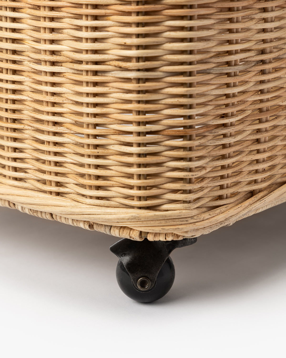 Close-up of the Chiara Laundry Basket in a natural wicker finish, featuring a black caster wheel attached to its bottom, placed on a white surface, by Vendixs