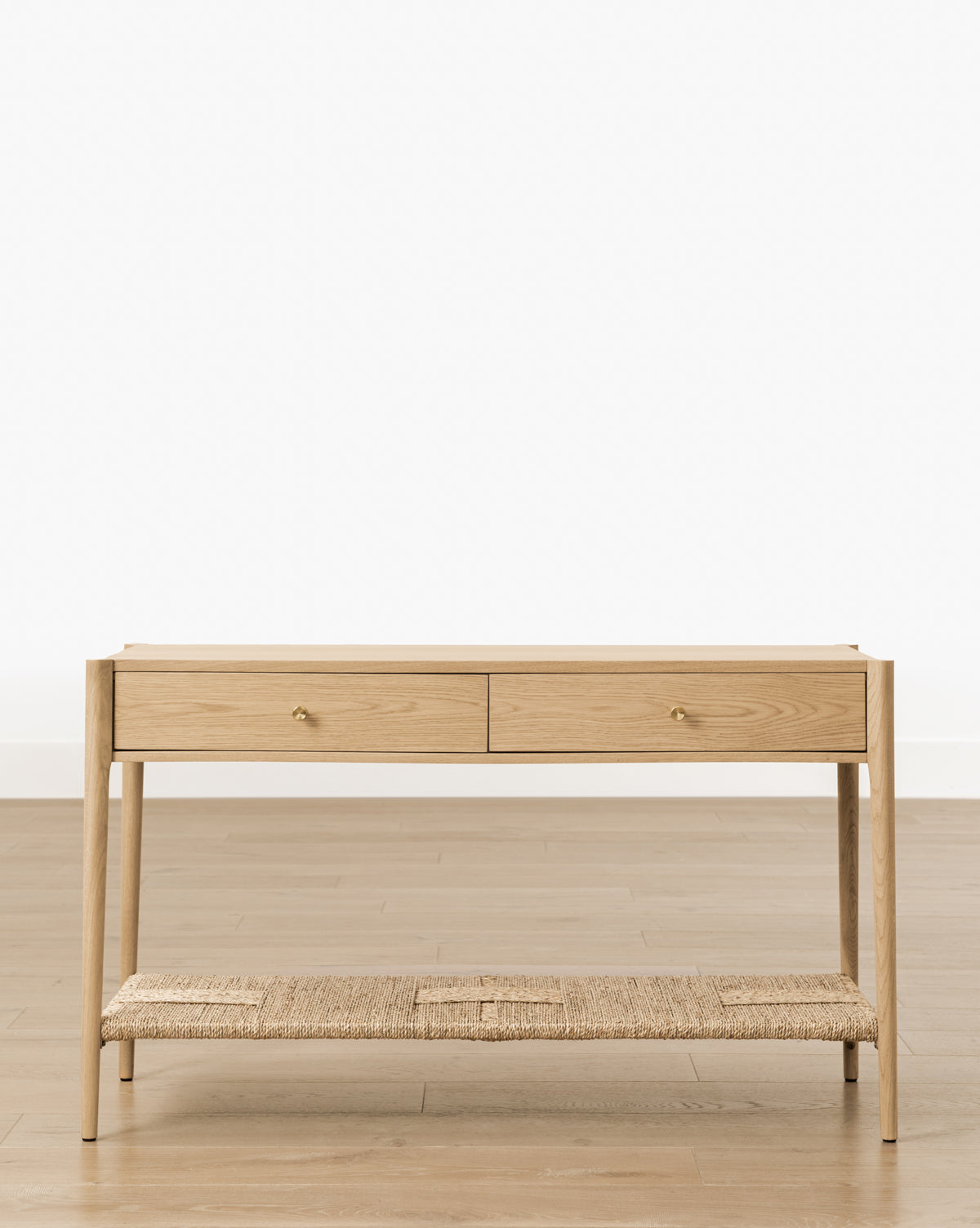 The Dana 2-Drawer Console features brass knobs and a woven lower shelf, ideal as an entryway accent, by Vendixs It stands on a light wood floor with a simple white background, offering both style and storage.