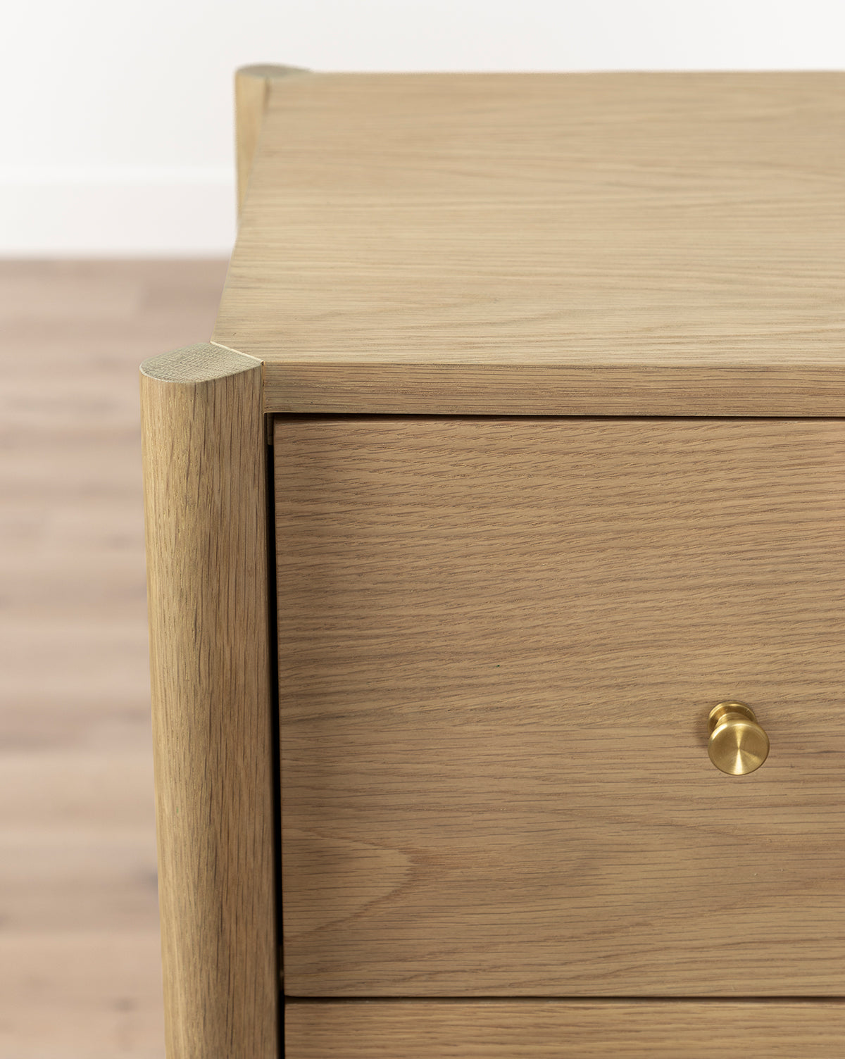 Close-up view of the Dana Tallboy Dresser in white oak, showcasing a single drawer with elegant brass hardware and smooth, natural wood grain. The background features a light wooden floor and a white wall, by McGee & Co.