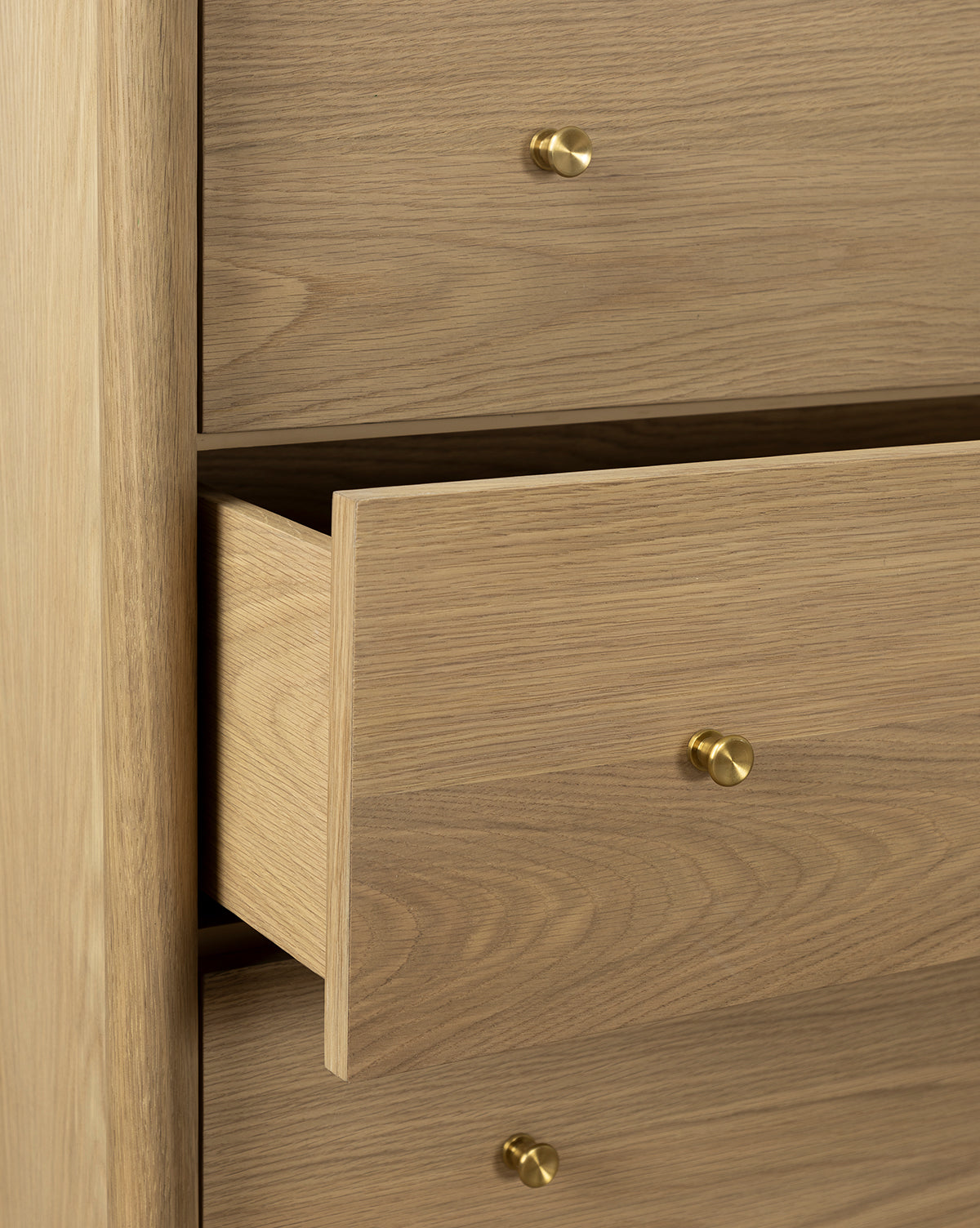 A close-up of the Dana Tallboy Dresser in white oak features round brass hardware, with one drawer partially open and two closed drawers above and below, by McGee & Co.