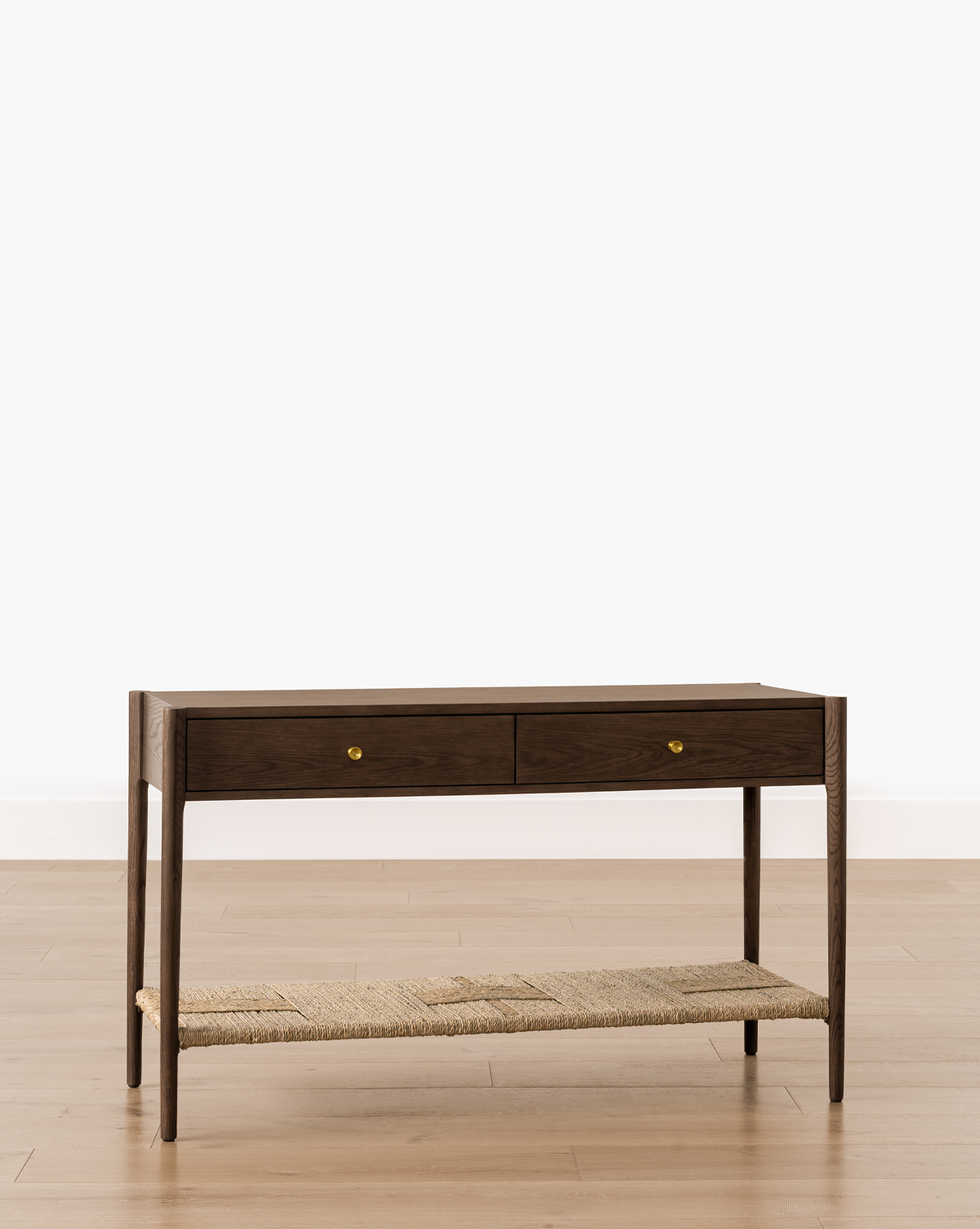 The Dana 2-Drawer Console, with brass knobs and a woven lower shelf in dark oak, brings timeless charm to your entryway when placed against a white wall and light wood floor, by Vendixs