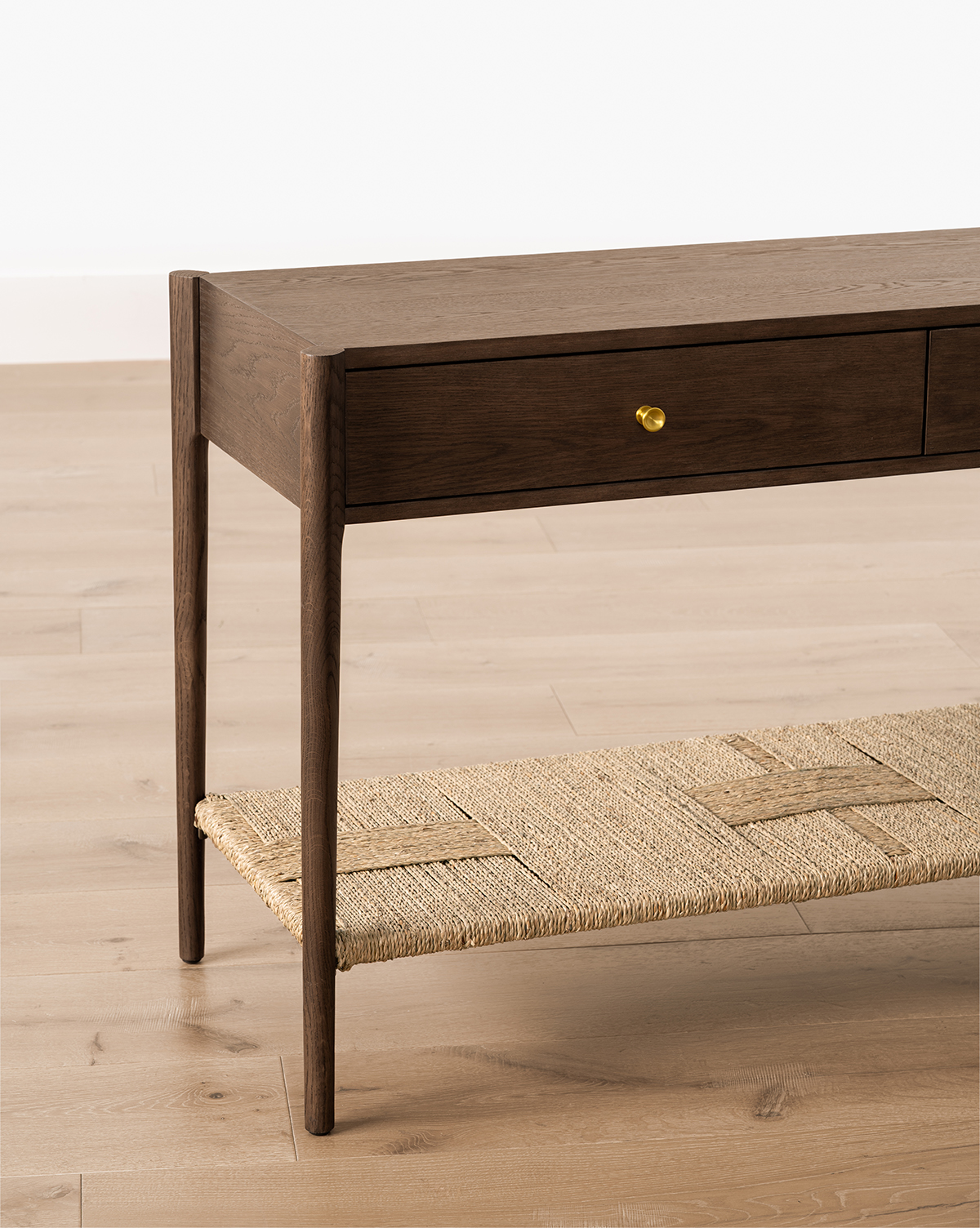 The Dana 2-Drawer Console features slim legs, brass knobs, and a woven rattan shelf to add charm to your entryway decor, set on a light wood floor against a white wall, by Vendixs