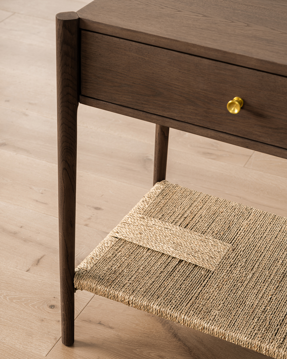 A close-up of the Dana 2-Drawer Console, showcasing its oak finish, round brass knobs, and woven lower shelf—ideal for entryway decor—set on a light wood floor, by Vendixs