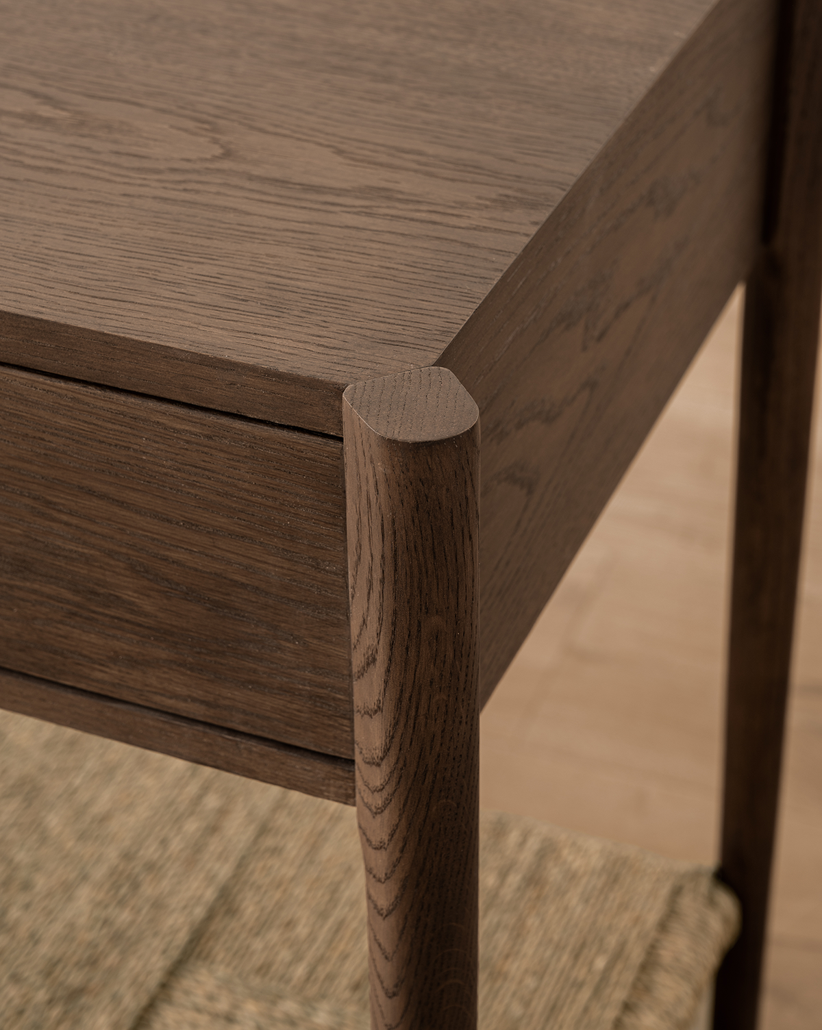 Close-up of the Dana 2-Drawer Console’s oak corner with rounded legs and visible wood grain, set above a woven mat on a light wood floor—ideal for adding style to any entryway, by Vendixs