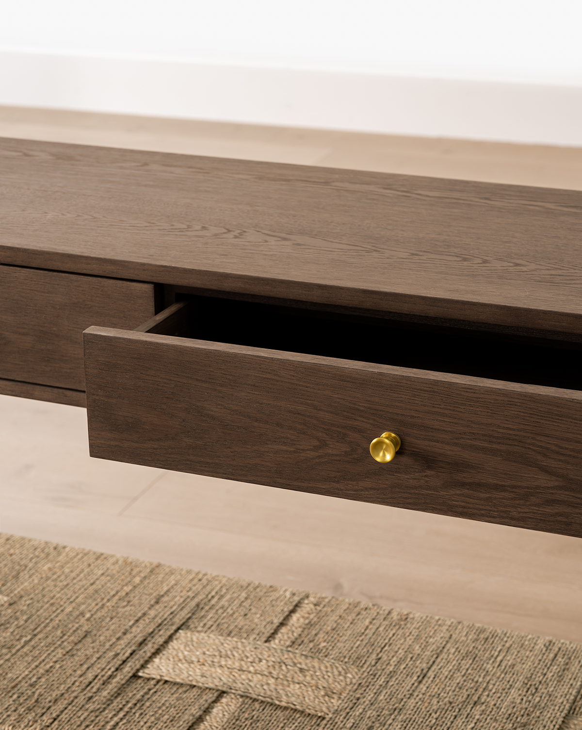 The Dana 2-Drawer Console in dark oak features a partially open drawer with a round gold knob, set on a light wood floor and woven beige rug—an elegant addition to entryway decor, by Vendixs