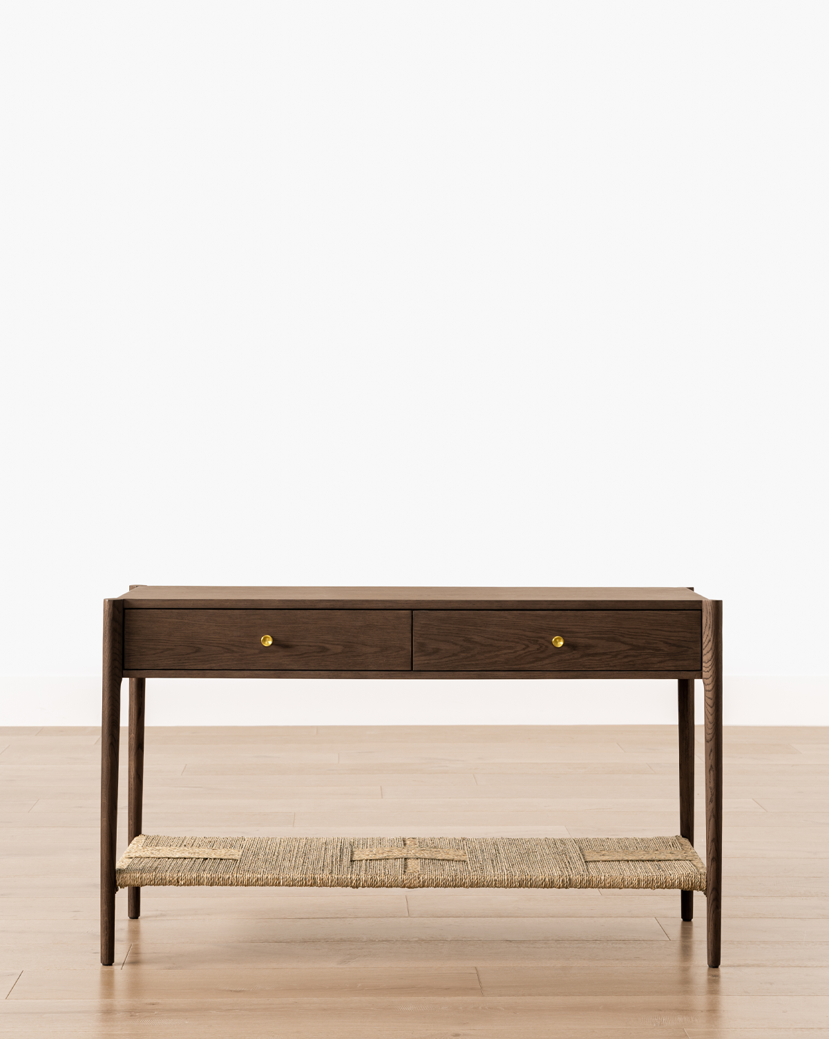 The Dana 2-Drawer Console, featuring gold knobs and a woven natural fiber lower shelf, stands on a light wood floor—perfect for entryway decor against a plain white wall, by Vendixs