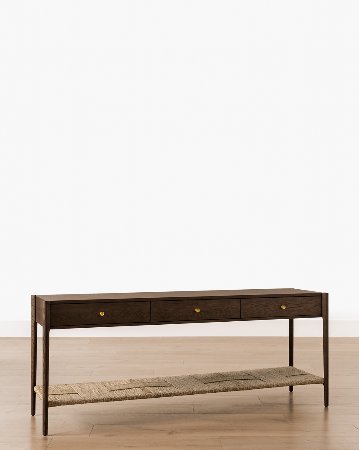 The Dana Walnut Oak Console features three small drawers with brass hardware and an open lower woven shelf, shown on a light wood floor against a plain white background, by Vendixs