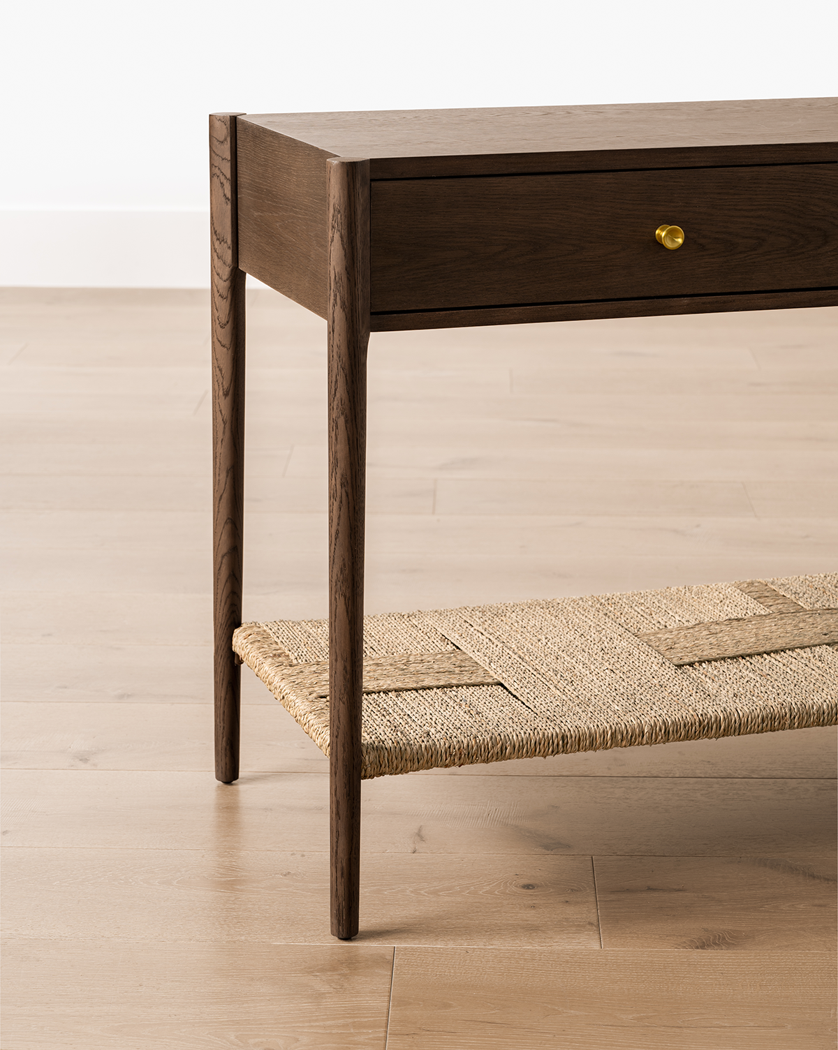 The Dana Walnut Oak Console, featuring slender legs, brass hardware on a single drawer, and a woven lower shelf, stands on a light wood floor against a white wall, by Vendixs